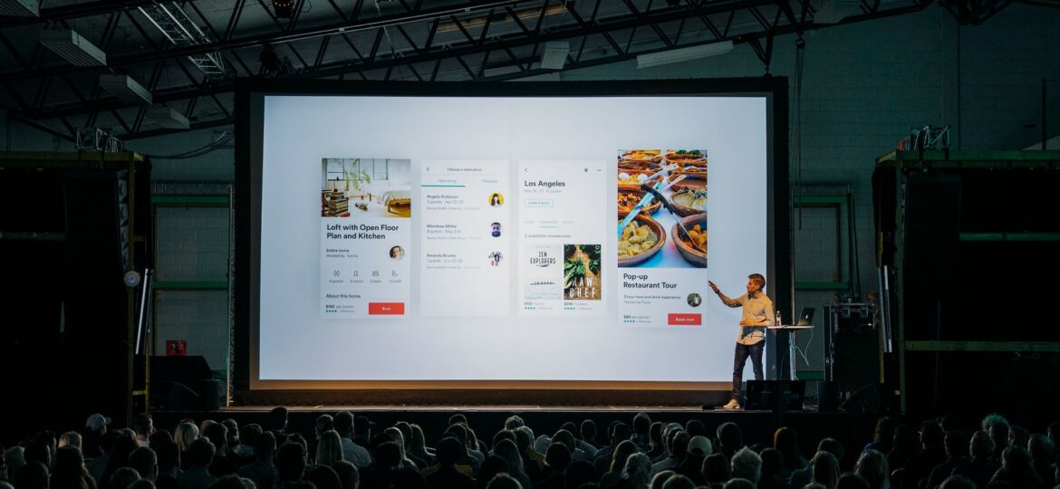 person discussing while standing in front of a large screen in front of people inside dim-lighted room